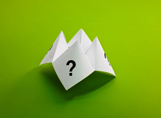 in Papier-Faltspiel mit aufgedruckten Fragezeichen liegt auf einem grünen Hintergrund – Symbol für Unentschlossenheit und Entscheidungsfindung.